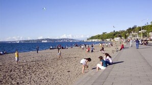 A beach with people walking and sitting, a clear sky, and a view of the sea and distant buildings.