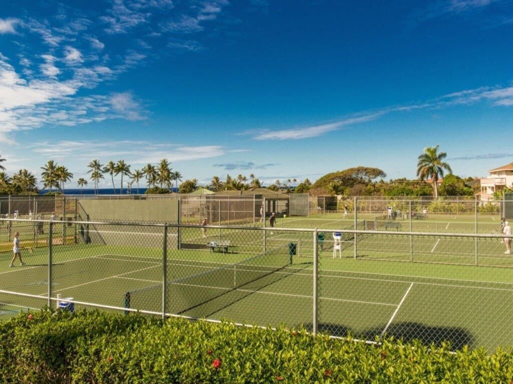 An outdoor tennis court with multiple courts, surrounded by greenery and palm trees.