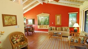 Uma sala de estar com parede vermelha, vigas de madeira no teto, ventilador de teto, um sofá, uma mesa de centro e um espaço de jantar com cadeiras e uma mesa.