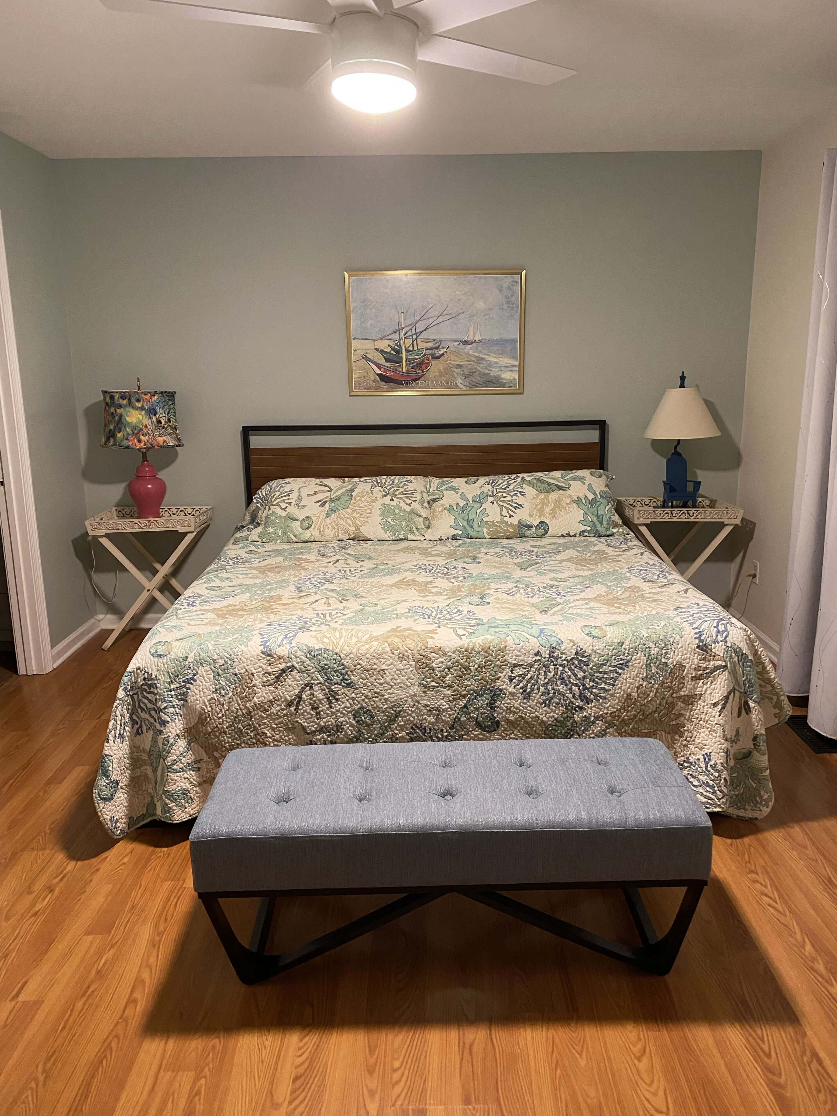 Un dormitorio con cama, banca, ventilador de techo, un cuadro y dos mesitas de noche con lámparas.