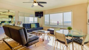 A living room with a large window, a ceiling fan, a flat-screen TV, and a seating area with a glass table and chairs. - Right on the Famous Mission Beach Boardwalk - Sleeps up to 10 on Ground Floor - Best location in Mission Beach (San Diego)