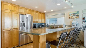 A modern kitchen with wooden cabinets, stainless steel appliances, a large island with bar stools, and a window providing natural light. - Right on the Famous Mission Beach Boardwalk - Sleeps up to 10 on Ground Floor - Best location in Mission Beach (San Diego)