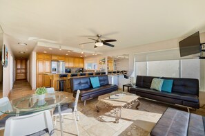 A modern kitchen with wooden cabinets, a dining area with a glass table, and a living room with a sofa and a TV.