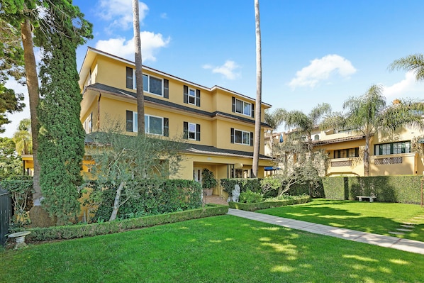 A two-story yellow building with a well-maintained lawn, a stone bench, and a pathway leading to the entrance.