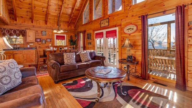 A cozy cabin interior with a wooden ceiling, a leather sofa, a round coffee table, and a large window with curtains.