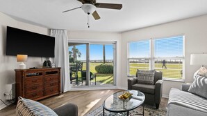 A living room with a ceiling fan, a flat-screen TV, a wooden cabinet, a sofa, a coffee table, and a view of a golf course through large windows.