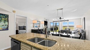 A modern kitchen with granite countertops and stainless steel sinks, a dining area with a glass table, and a living room with a sofa and a TV.