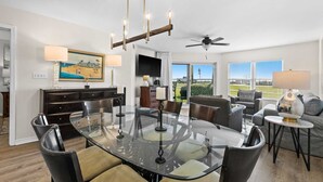 A spacious dining area with a glass table and chairs, a ceiling fan, and a view of the outdoors.