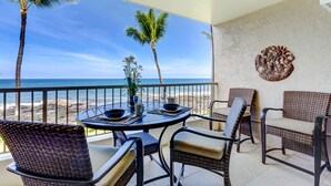 A balcony with wicker chairs and a table, overlooking a beach and ocean. - Oceanfront Condo, 5G Wi-Fi, King Bed, Hot Tub/Pool, Free Parking (Kailua-Kona)