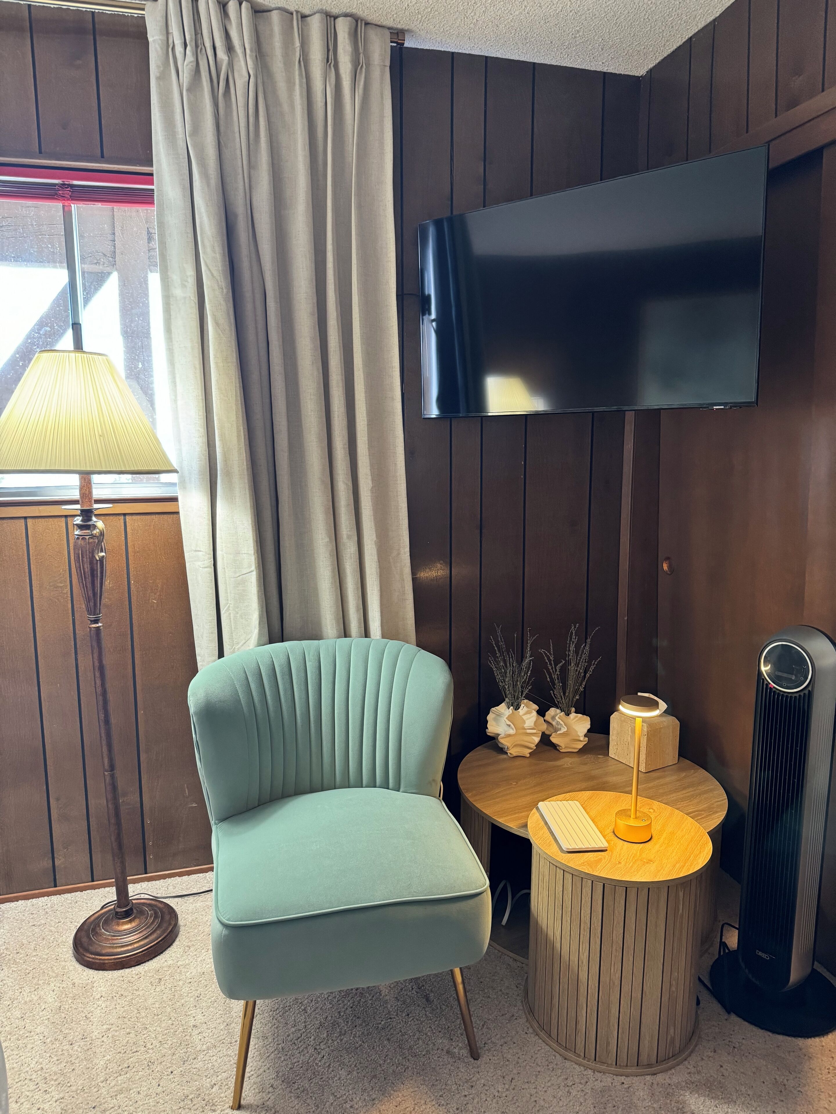 Een moderne kamer met een blauwgroene fauteuil, een ronde houten tafel met een lamp, een flatscreen televisie en een radiator.