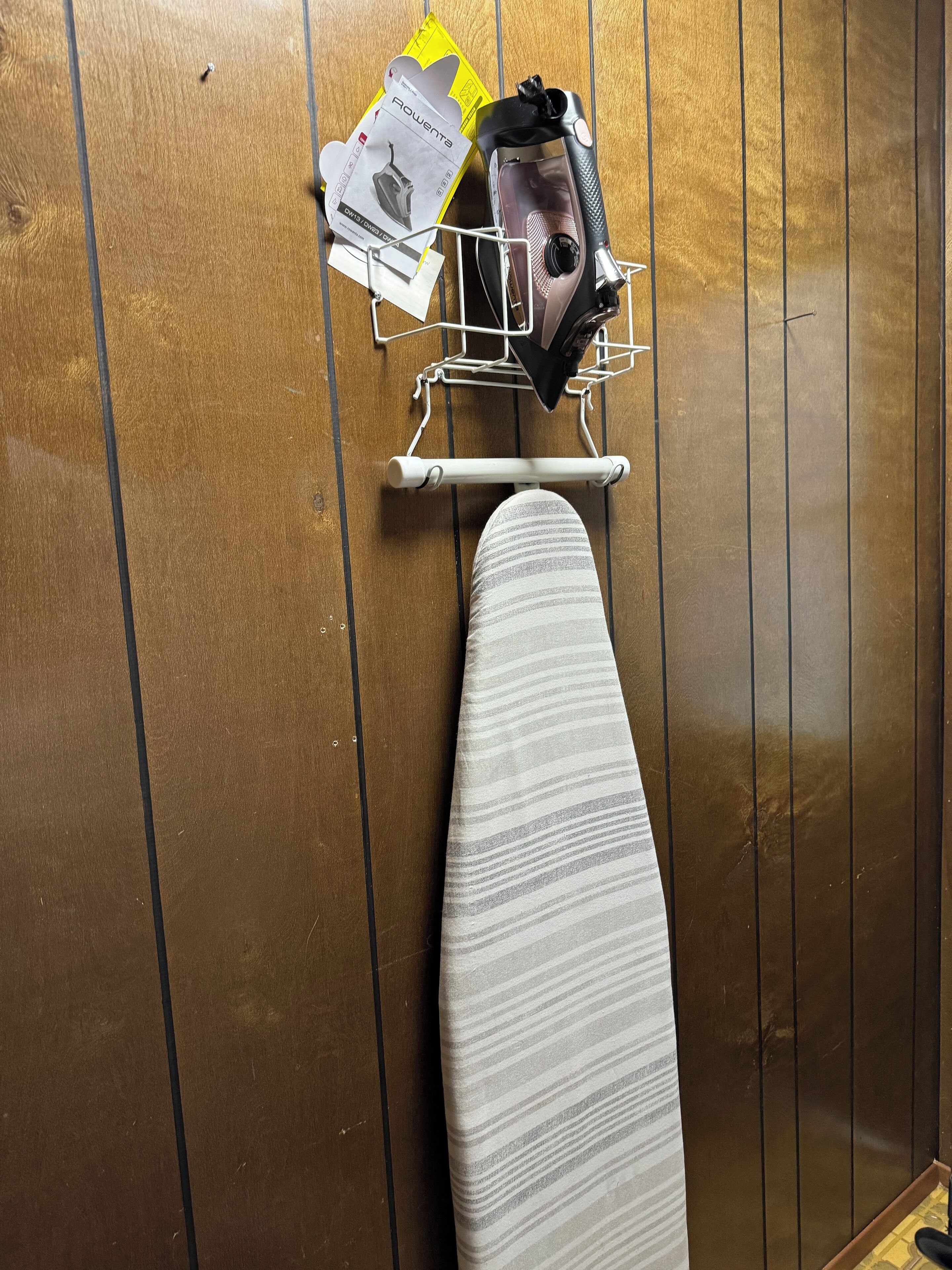 Un mur recouvert de panneaux de bois, sur lequel on aperçoit une planche à repasser blanche et un étagère métallique portant un sac et des papiers.