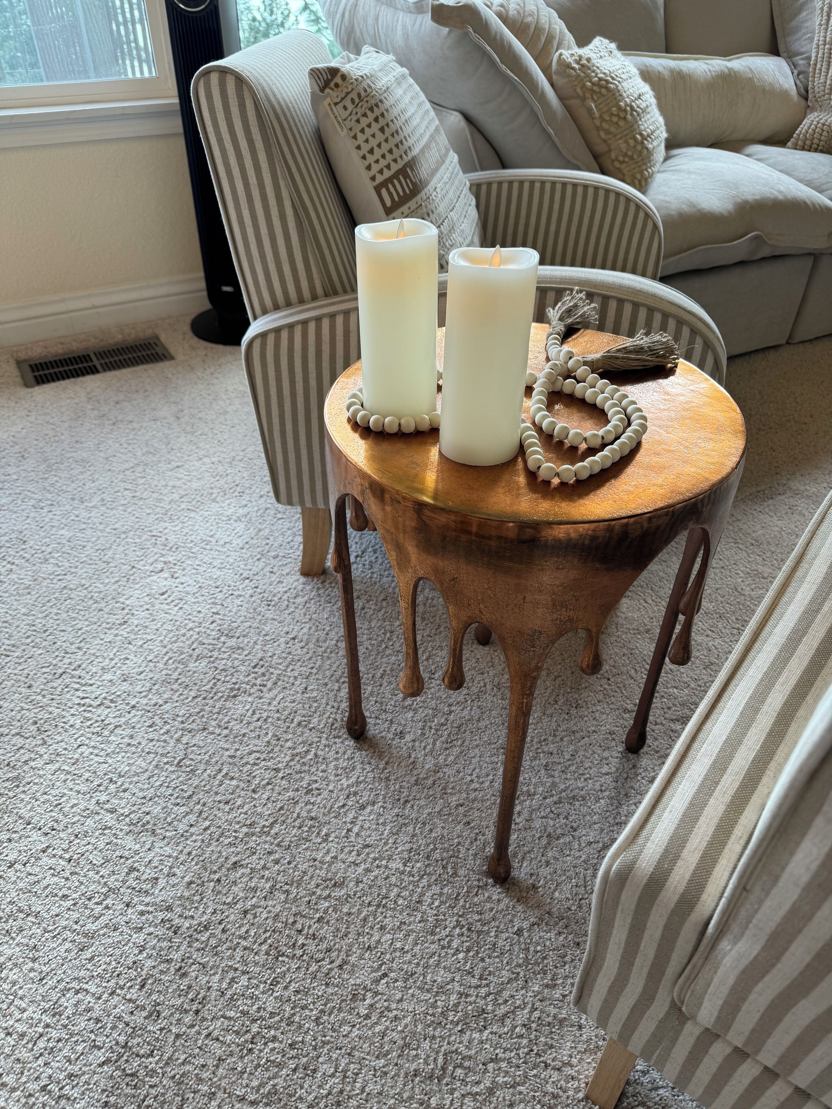 A cozy living room with a striped armchair, a round wooden coffee table with candles and a beaded necklace, and a beige sofa.