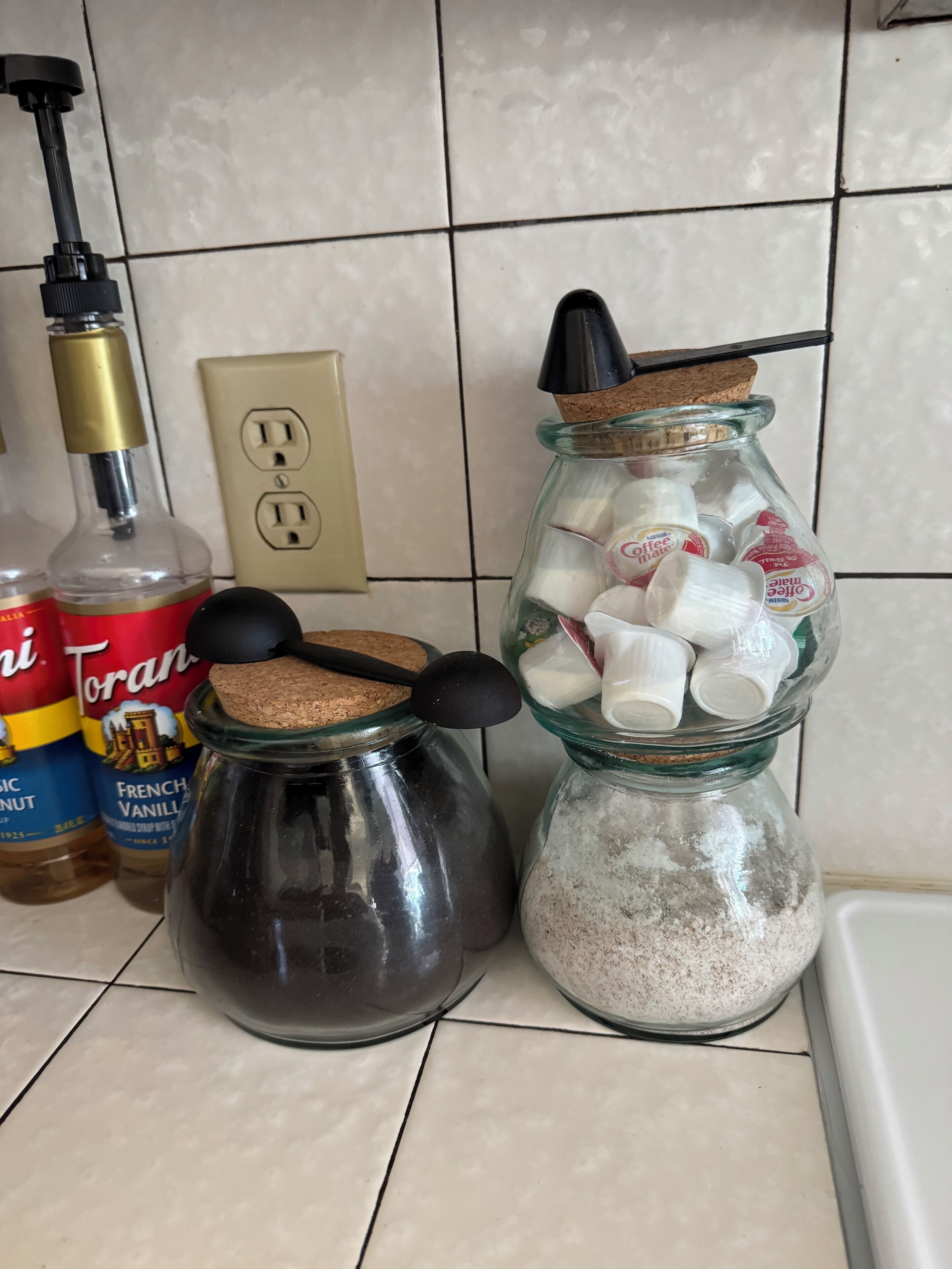 A kitchen counter with various containers, including a jar with a cork stopper and a metal lid, a jar with a black lid, and a bottle of French vanilla syrup.