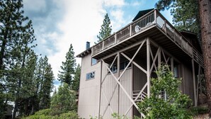 Una cabaña con terraza de madera y estructura metálica, rodeada de un bosque frondoso.