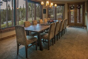 Un espace repas avec une grande table en bois et des chaises à motifs, de grandes fenêtres donnant sur la forêt et un lustre.