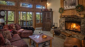 A cozy cabin interior with a fireplace, wooden walls, and large windows overlooking a forest. - SmallGroup-SmallRate! Great Layout/Views/HotTub/PoolTable/Grills/Ski In Ski Out! (Breckenridge)