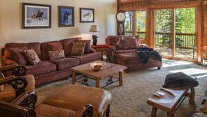 A cozy living room with a large sofa set, a coffee table, and a bench. There are three framed pictures on the wall, a clock, and a window with a view of trees. - SmallGroup-SmallRate! Great Layout/Views/HotTub/PoolTable/Grills/Ski In Ski Out! (Breckenridge)