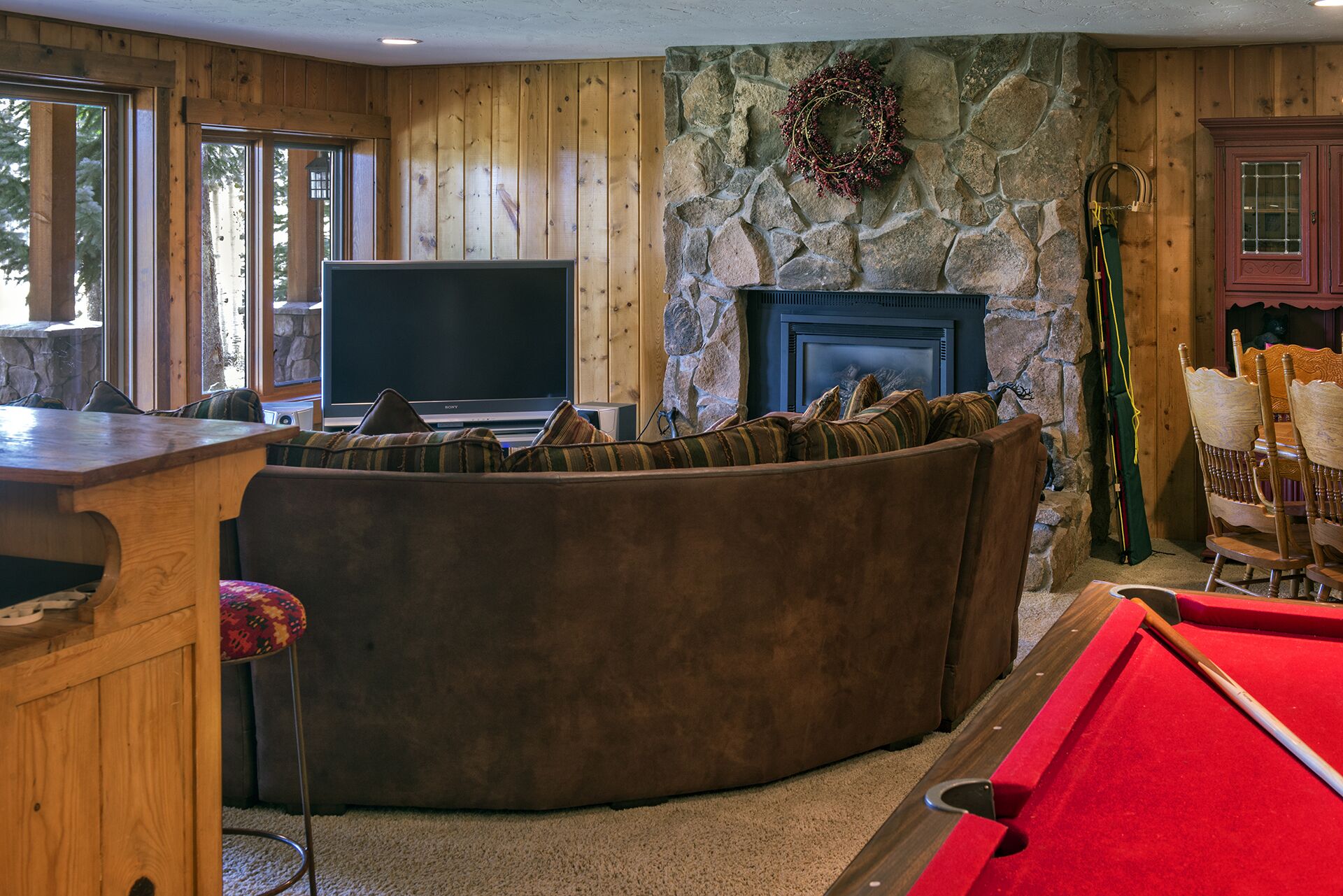 A cozy living room with a stone fireplace, a flat-screen TV, a pool table, and wooden paneling.