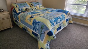 A bedroom with a bed covered in a tropical-themed quilt, a wooden headboard, a nightstand with a clock, and a window with blinds.