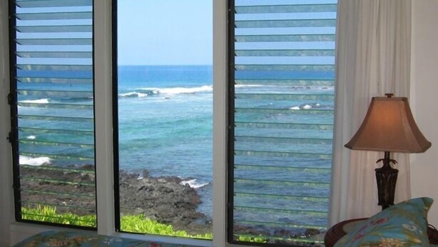 A room with a bed, a lamp, and a view of the ocean through a window with blinds. - Beautiful Ocean/Beachfront Property and A Private Back Yard!  AC in both BDRMS! (Kailua-Kona)