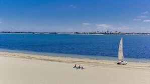 A beach with a sailboat near the shore, people relaxing on the sand, and a clear view of the ocean and distant shoreline. - Bayfront - Mission Bay on Sand Panoramic View -Upscale (San Diego)