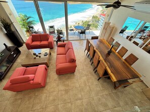 A spacious living room with a large window overlooking a beach, furnished with red sofas and a wooden dining table. - Casa Spadaro - Luxury Amara Beachfront Penthouse with private rooftop terrace (Ixtapa Zihuatanejo)