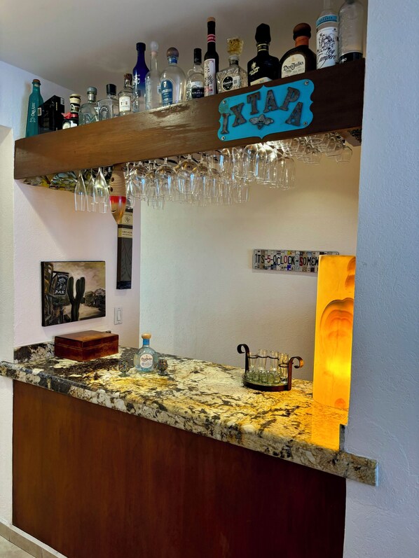 A bar area with a wooden shelf holding various liquor bottles and glasses. - Casa Spadaro - Luxury Amara Beachfront Penthouse with private rooftop terrace (Ixtapa Zihuatanejo)