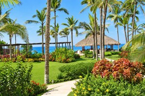 A tropical garden with palm trees, a thatched-roof structure, and a view of the ocean. - Casa Spadaro - Luxury Amara Beachfront Penthouse with private rooftop terrace (Ixtapa Zihuatanejo)