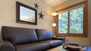Un salon moderne avec un canapé en cuir foncé, une table d’appoint en bois sur laquelle repose un plateau, une lampe sur pied et une grande fenêtre donnant sur la forêt.