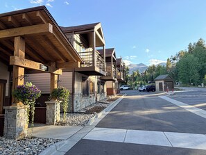 A multi-story building with a stone facade, wooden balconies, and a parking area with vehicles.