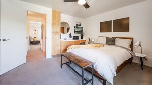Un dormitorio con cama, una banca, una mesita de noche con lámpara, ventilador de techo y obras de arte en la pared.