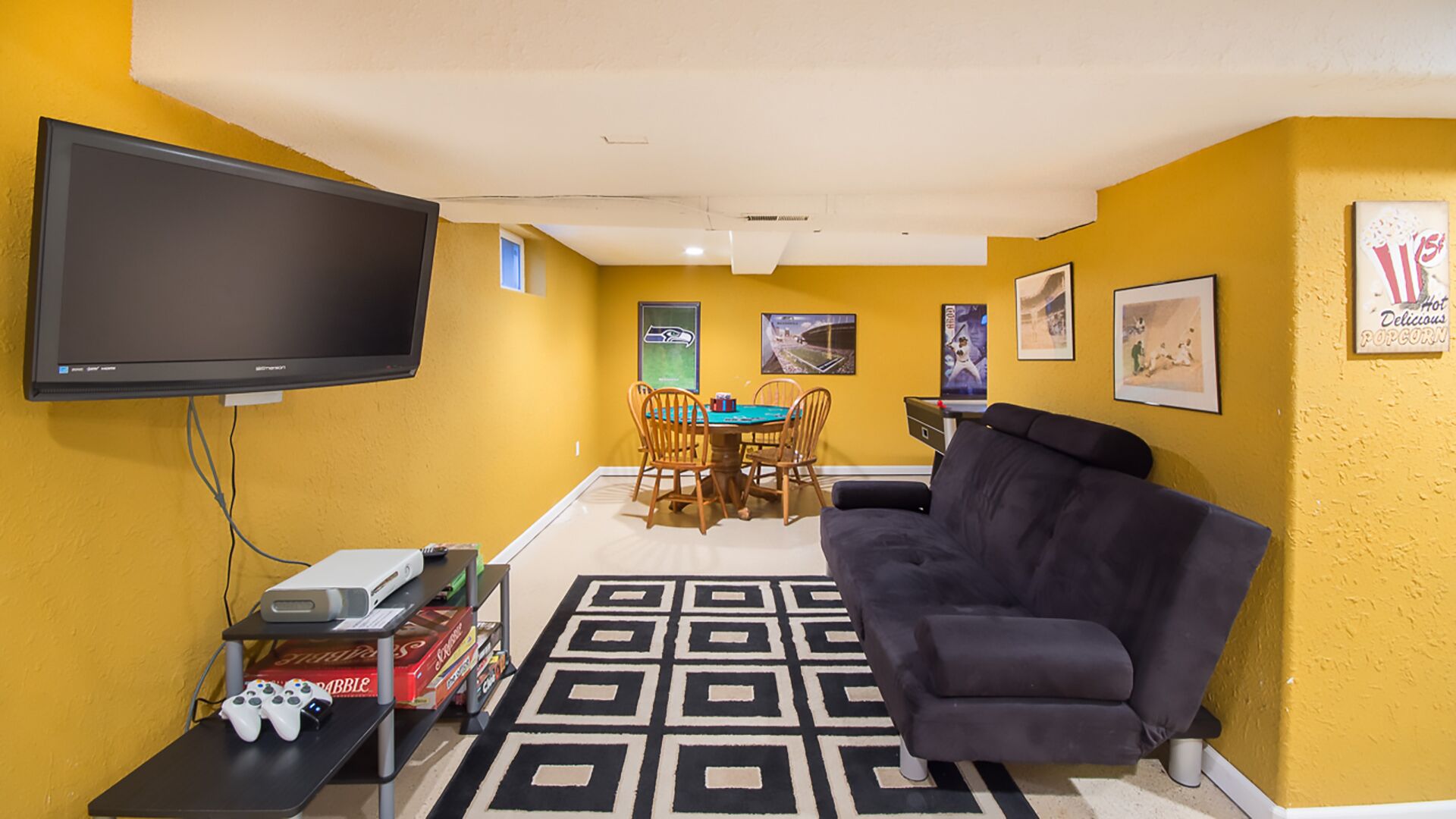 Un espacio habitable compacto con una televisión de pantalla plana, una consola de videojuegos, un sofá, una mesa de comedor con sillas y una alfombra estampada.
