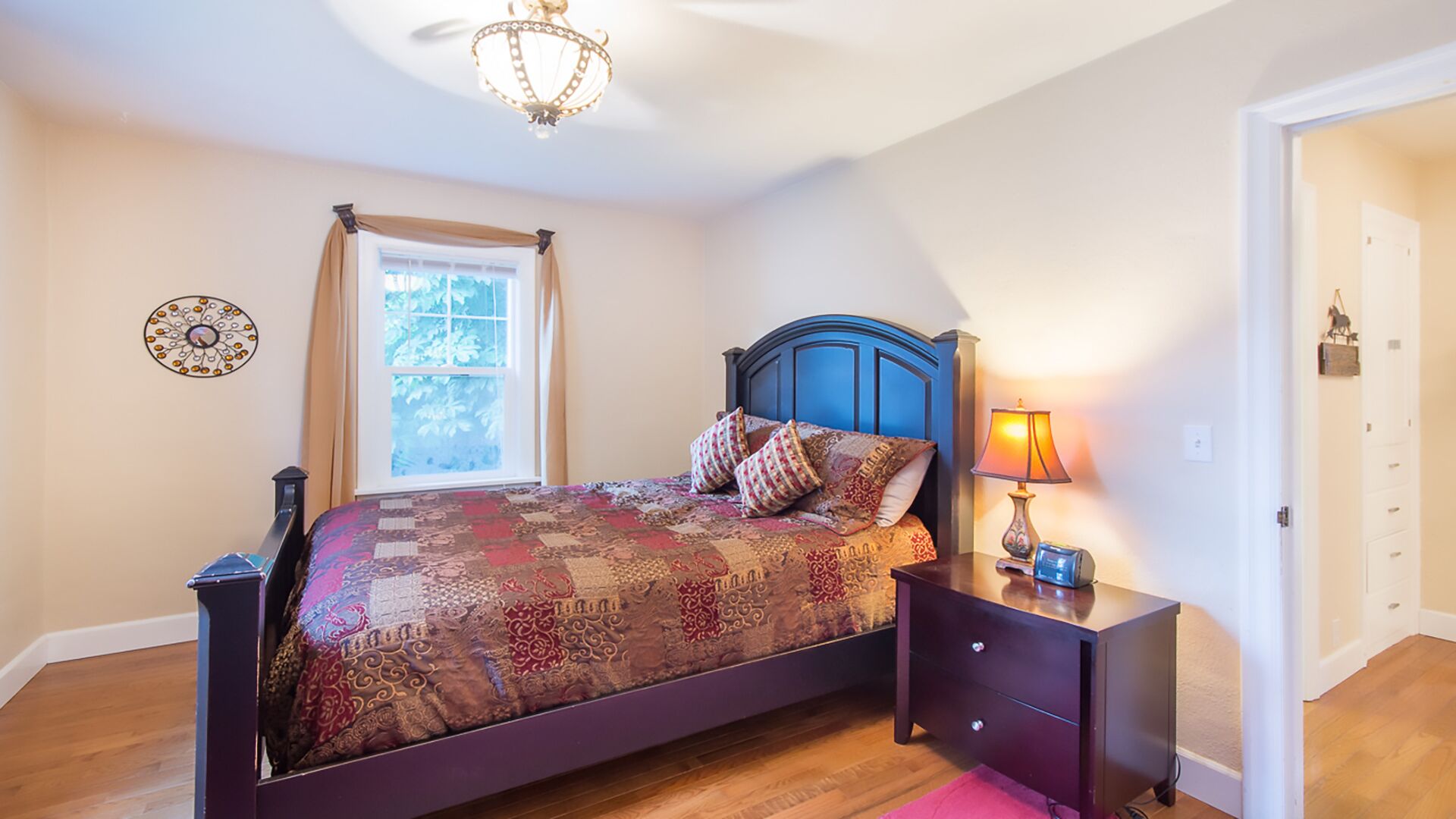 A bedroom with a bed, a nightstand, a lamp, a window with curtains, and a chandelier.
