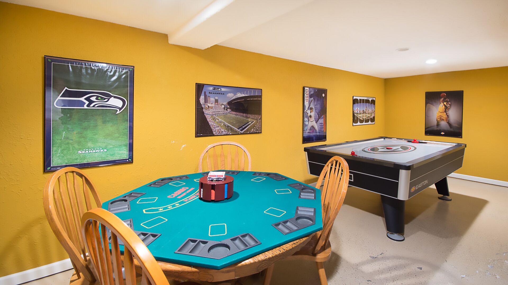 A room with a pool table, a foosball table, and a table with poker chips. There are posters on the walls, including one with a football stadium and another with a basketball player.
