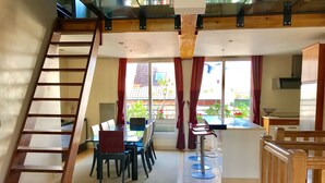 A spacious room with a wooden staircase, a dining area with a glass table, and a kitchen area with a bar counter.