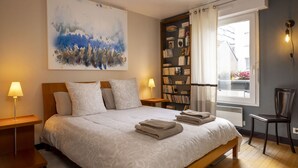 A neatly made bed with a large abstract painting above it, a bookshelf with books and decorative items, and a window with curtains.