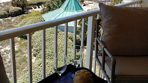 A balcony with a view of the beach, a green-roofed structure, and a chair with a cushion.