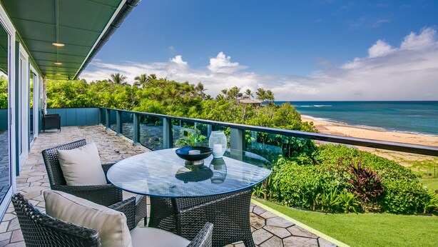 A glass-top table with wicker chairs and a bowl, overlooking a beach with palm trees and a clear sky. - Hale Pono- Oceanfront, private, gated, great views, AC, large property  (Haleiwa)