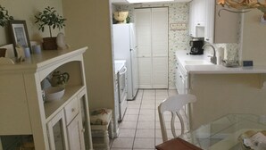 A compact kitchen with a refrigerator, a dining table with chairs, a small cabinet, and a potted plant. - St. Simons Island Beach Club Condo #126 (St. Simons Island)