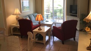 A living room with a sofa, two armchairs, a coffee table, a lamp, a TV, and a balcony with a view of greenery. - St. Simons Island Beach Club Condo #126 (St. Simons Island)
