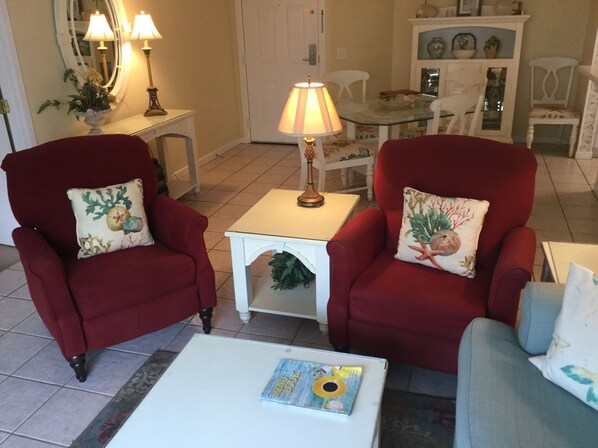 A living room with two red armchairs, a white coffee table, and a glass-top table with chairs. - St. Simons Island Beach Club Condo #126 (St. Simons Island)