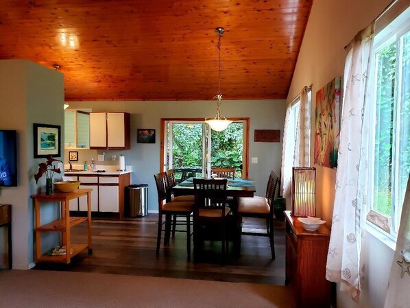 A spacious living area with a wooden ceiling, a dining table with chairs, a kitchen area, and large windows with curtains.