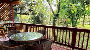 Una terraza de madera con una mesa de vidrio y sillas de mimbre, rodeada por una barandilla roja y abundante vegetación.