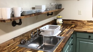 Una cucina con piano di lavoro in legno, lavello in acciaio inox e mobili verdi. Si vedono mensole con piatti e bicchieri, e un vaso in ceramica a tema frutta appeso al muro.