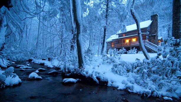 Una cabaña de troncos con chimenea de piedra, rodeada de árboles cubiertos de nieve y un arroyo.