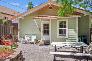 A small, single-story house with a front porch, a door, and a window. There are two white chairs and a table on the porch.