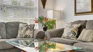 Una sala de estar con un sofá gris, cojines estampados, una mesa de centro de vidrio, un jarrón con plantas, una lámpara y un espejo enmarcado en la pared.