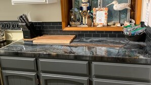 A kitchen with dark gray cabinets, a black marble countertop, and a window with a view of greenery. - Beverly beach delight (Newport)