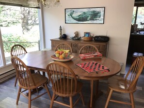 Un comedor con una mesa de madera, sillas, un frutero, un tablero de ajedrez y un juego de damas.