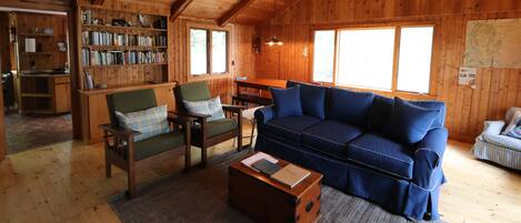 A cozy cabin interior with a wooden ceiling, a blue sofa, a green armchair, a wooden coffee table, and a bookshelf filled with books.